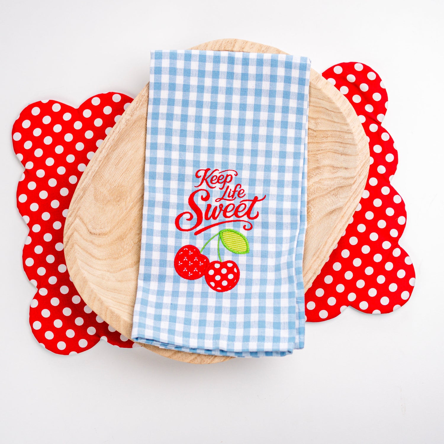 Blue checkered towel with 'Keep Life Sweet' text and cherry design on a wooden plate with red polka dot coasters.