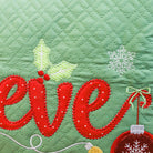 Close-up of a green quilted fabric with embroidered holiday lettering reading "eve" in red, white snowflakes, holly leaves, and a red ornament.