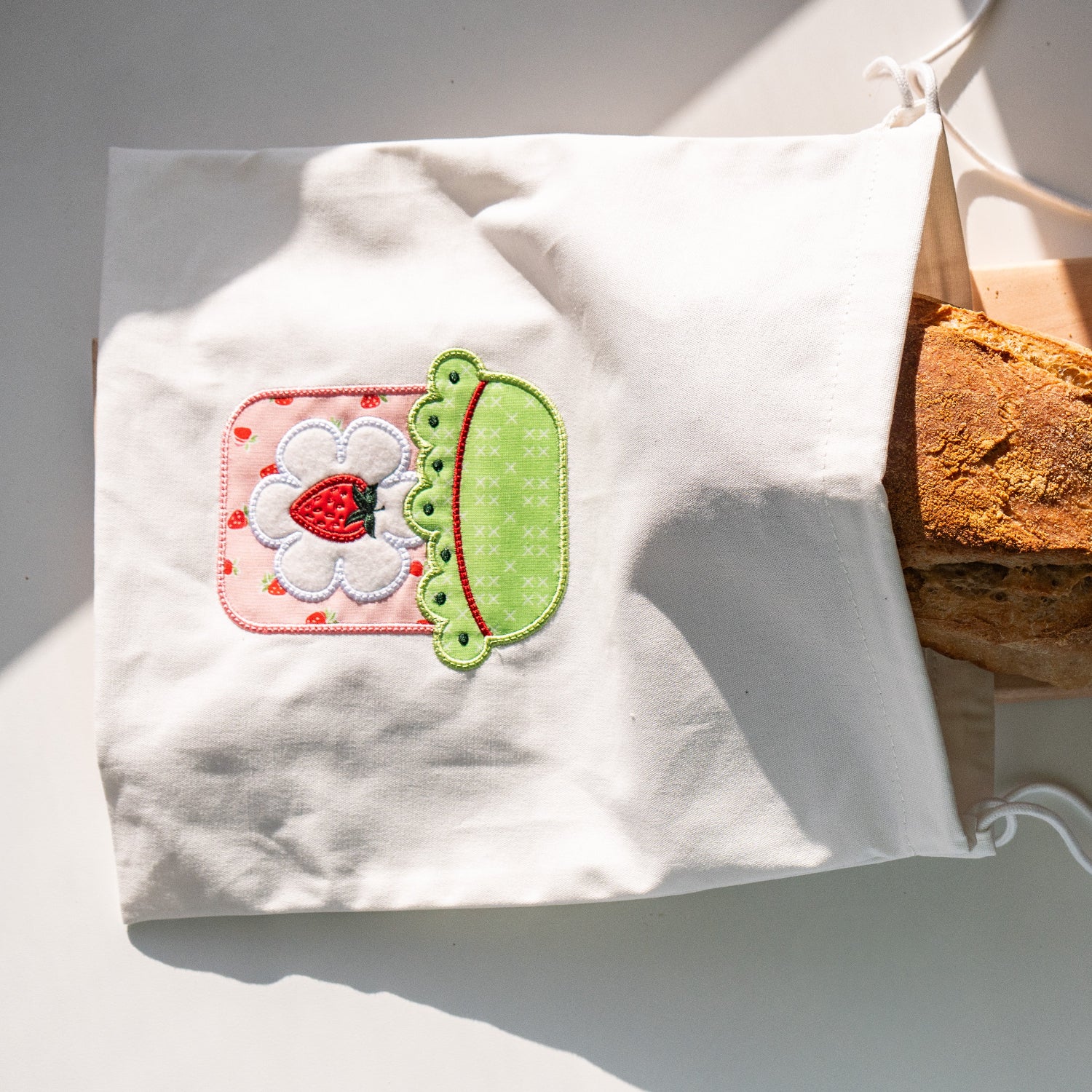 White tote bag with a decorative patch containing a strawberry and kiwi, filled with bread on a white surface.