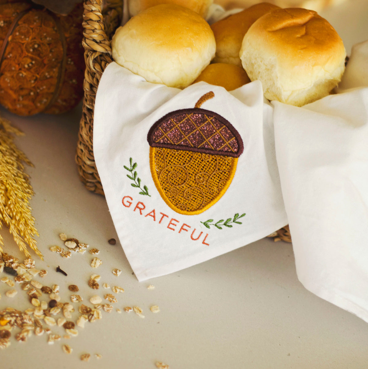 A white fabric napkin with an embroidered acorn design and the word 'GRATEFUL' in red, styled with bread rolls and autumn-themed props on a tabletop.