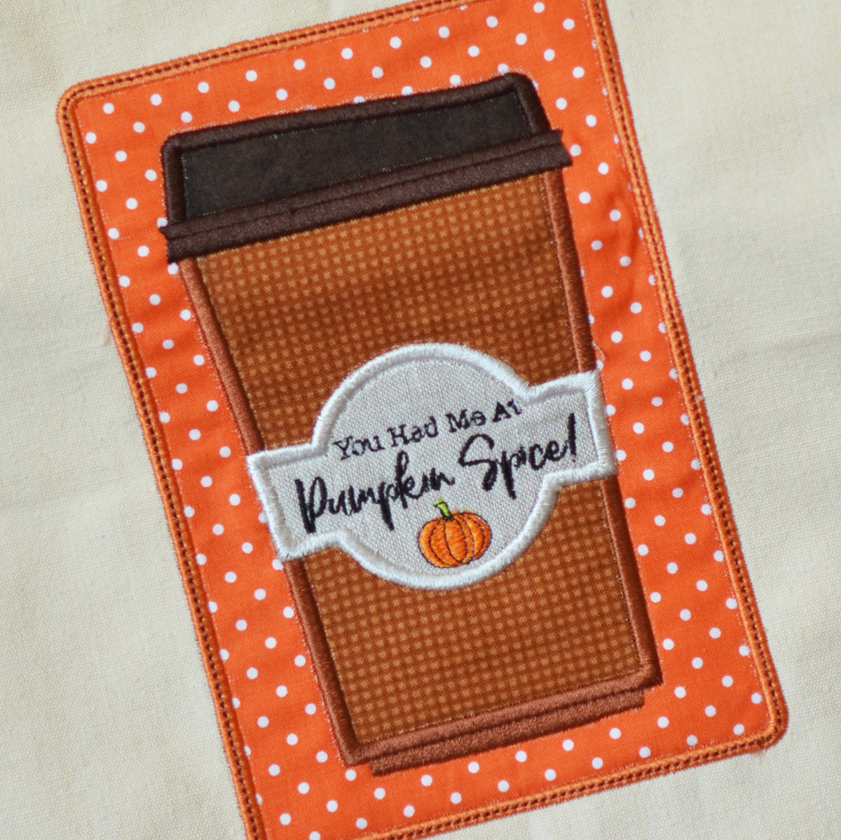 An embroidered applique design of a coffee cup with a brown polka-dot body, a dark brown lid, and a white circular label reading 'You Had Me At Pumpkin Spice!' with a small pumpkin graphic, set on an orange and white polka-dot background.