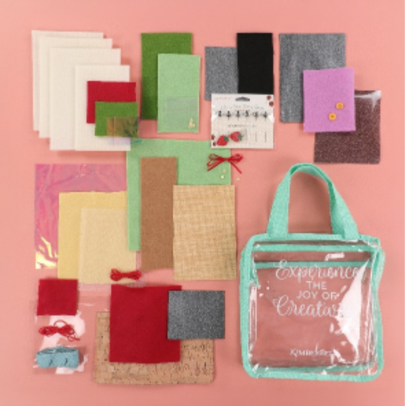 A flat-lay display of a craft kit featuring various colorful felt sheets, fabric swatches, and a clear tote bag with mint green handles and trim on a solid pink background.
