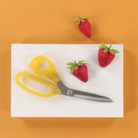 A pair of yellow-handled scissors with silver blades rests on a white rectangular board, with three small red strawberries placed around it on a solid orange background.