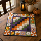 A square quilted table runner with a Halloween theme sits on a wooden dining table. The runner features a central checkerboard pattern in purple, orange, green, black, and white, surrounded by a border of autumn-themed patches including pumpkins, corn, flowers, and text like 'SWEET SPOT', 'HAPPY HALLOWEEN', 'ROO', and 'BE KIND'.