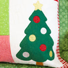 A close-up of a handmade Christmas-themed patchwork pillow featuring an applique green Christmas tree with red, gold, and white sequin ornaments on a white background, surrounded by red, green, and pink patterned fabric panels.