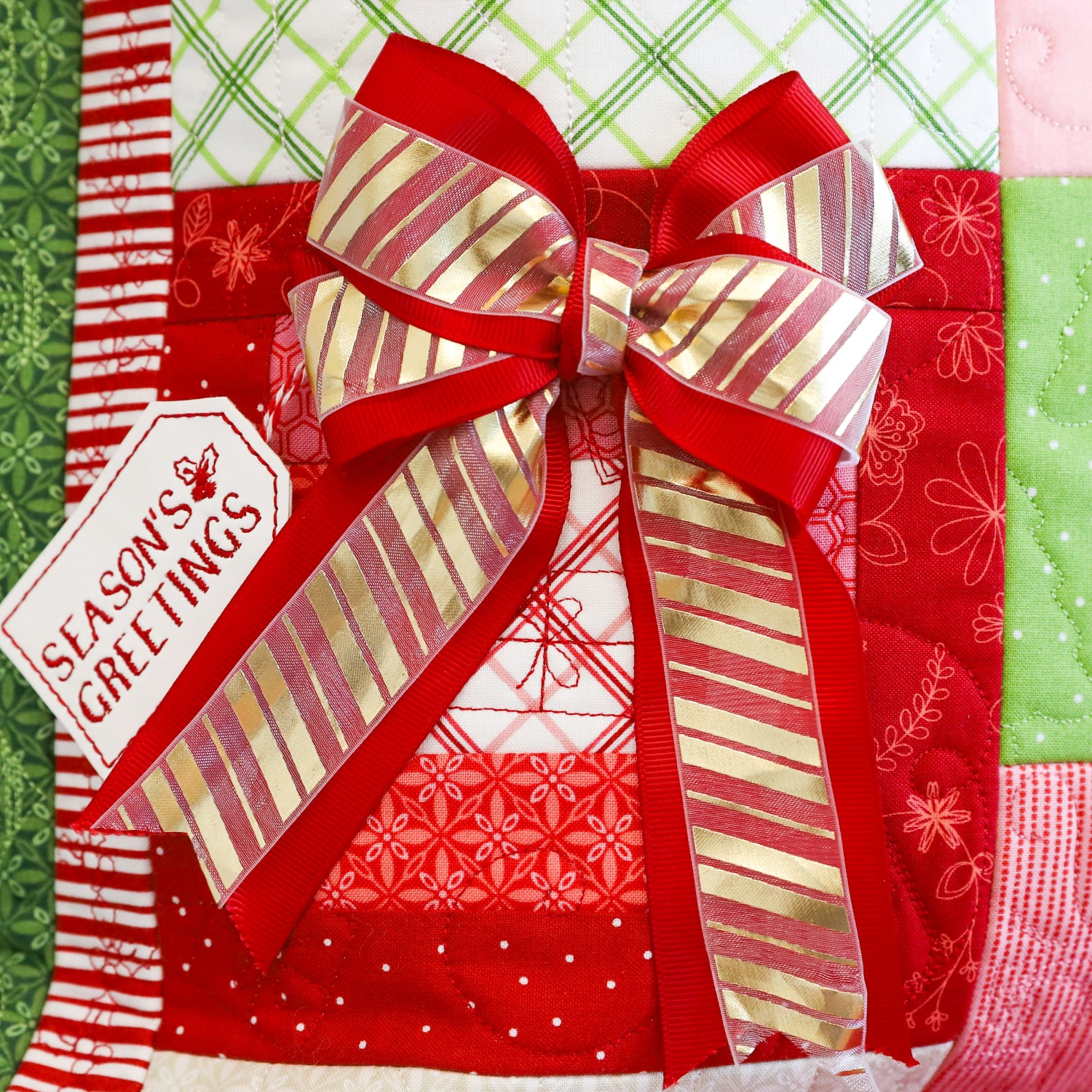 A close-up of a quilted fabric surface with a large red and gold striped ribbon bow, a white paper tag reading 'Seasons & Greetings,' and patchwork-style holiday fabric panels in red, green, pink, and white.