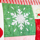 Close-up of a quilted fabric panel with a green square featuring a white embroidered snowflake, surrounded by red, white, and pink holiday-themed fabric sections.