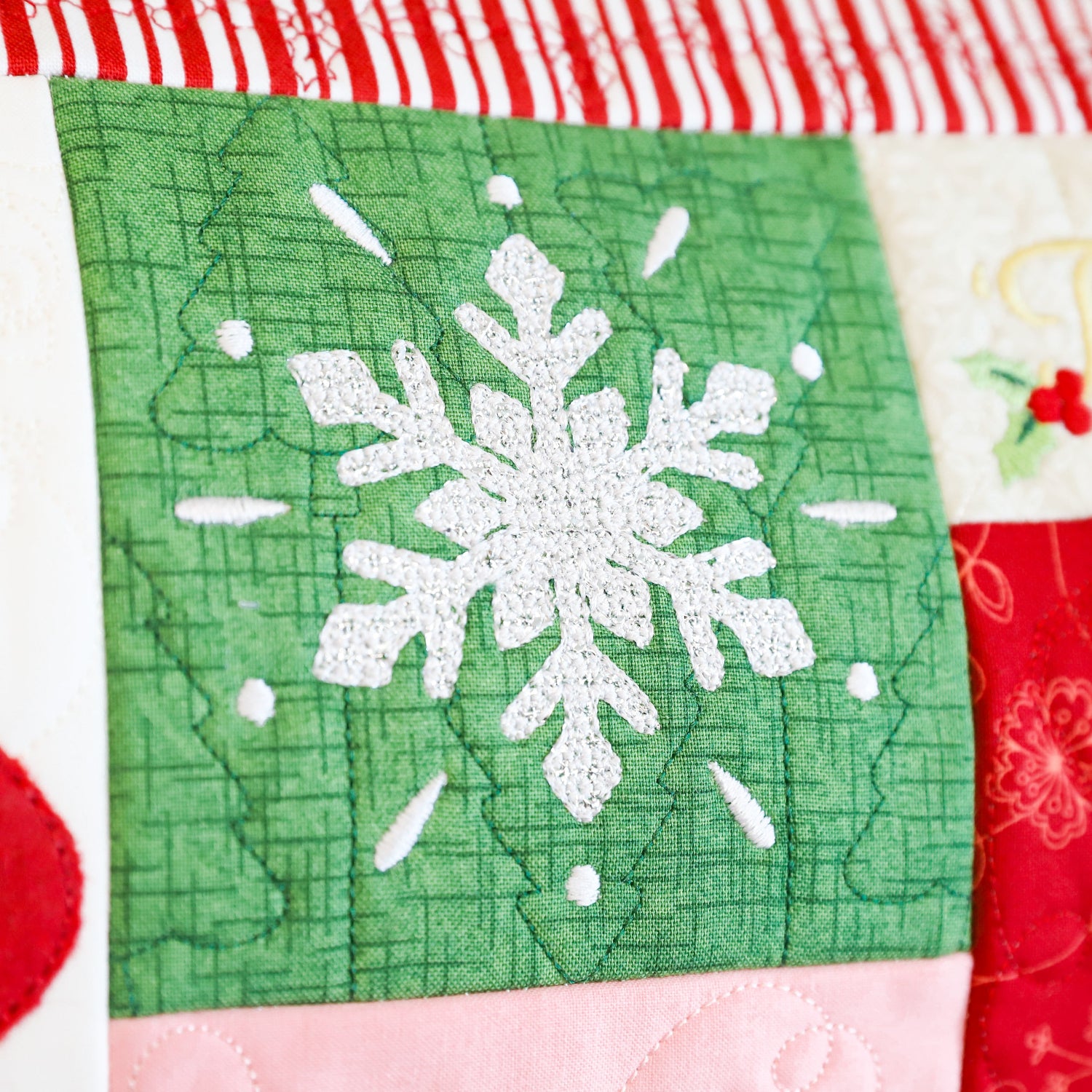 Close-up of a quilted fabric panel with a green square featuring a white embroidered snowflake, surrounded by red, white, and pink holiday-themed fabric sections.
