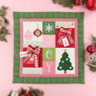 A square quilted Christmas wall hanging or placemat with a green patterned border and a central patchwork design featuring holiday ornaments, a snowflake, the letters J, O, Y, a Christmas tree, and gift boxes with red bows, styled on a pink background with evergreen branches and ornaments.