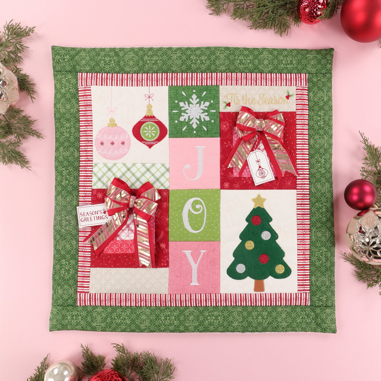 A square quilted Christmas wall hanging or placemat with a green patterned border and a central patchwork design featuring holiday ornaments, a snowflake, the letters J, O, Y, a Christmas tree, and gift boxes with red bows, styled on a pink background with evergreen branches and ornaments.