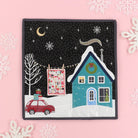 A square quilted fabric art piece featuring a festive Christmas night scene, displayed on a light pink background decorated with white snowflakes. The quilt depicts a teal house with snow on the roof, a red car with Christmas gifts, a bare tree, and a quilt hanging on a clothesline under a starry black sky.