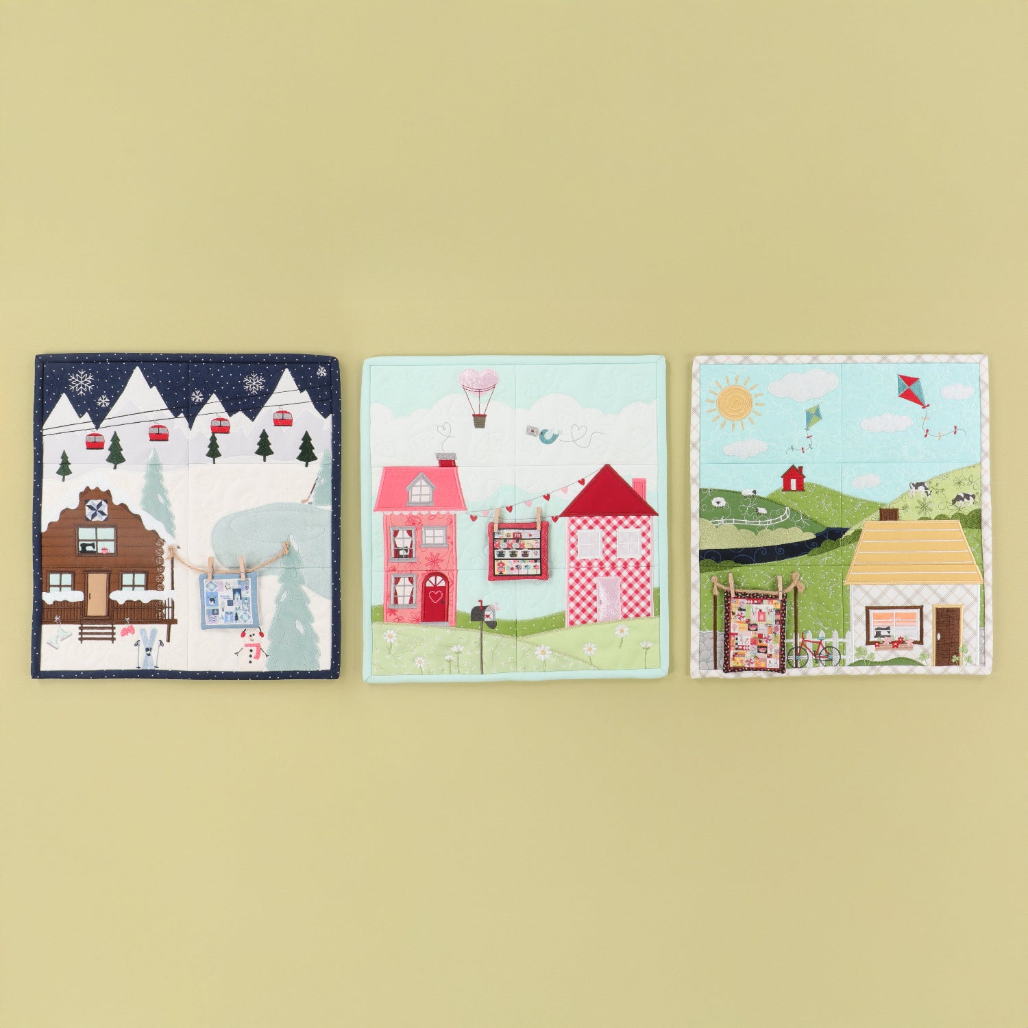 Three small square embroidered art cards or patches with whimsical village scenes, displayed side by side on a solid pale yellow background.
