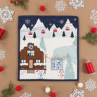 A square quilted fabric Advent calendar page with a winter mountain cabin scene, styled in a flat lay on a tan background with white snowflake cutouts, red pom-poms, green pine branches, and red spools of thread.