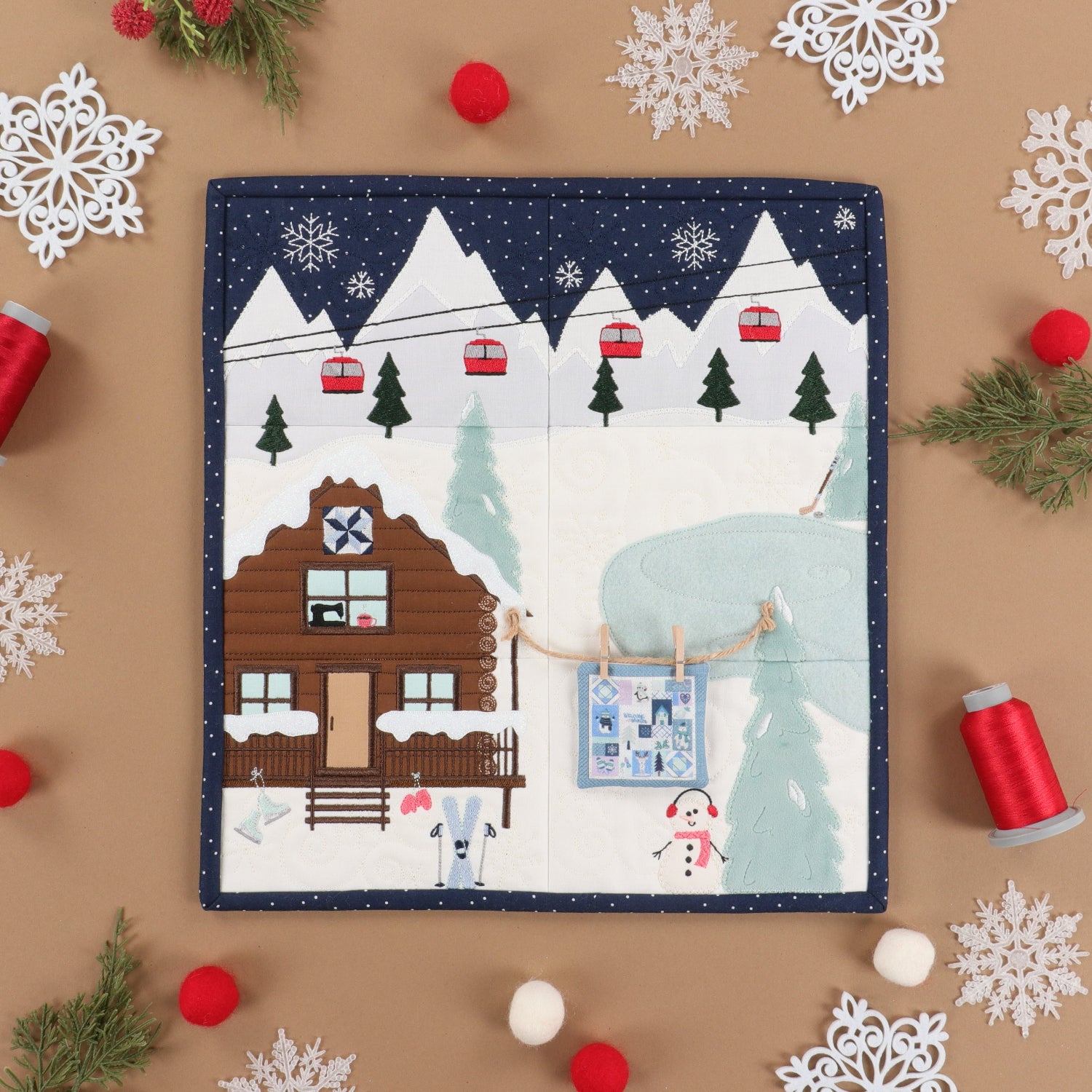A square quilted fabric Advent calendar page with a winter mountain cabin scene, styled in a flat lay on a tan background with white snowflake cutouts, red pom-poms, green pine branches, and red spools of thread.