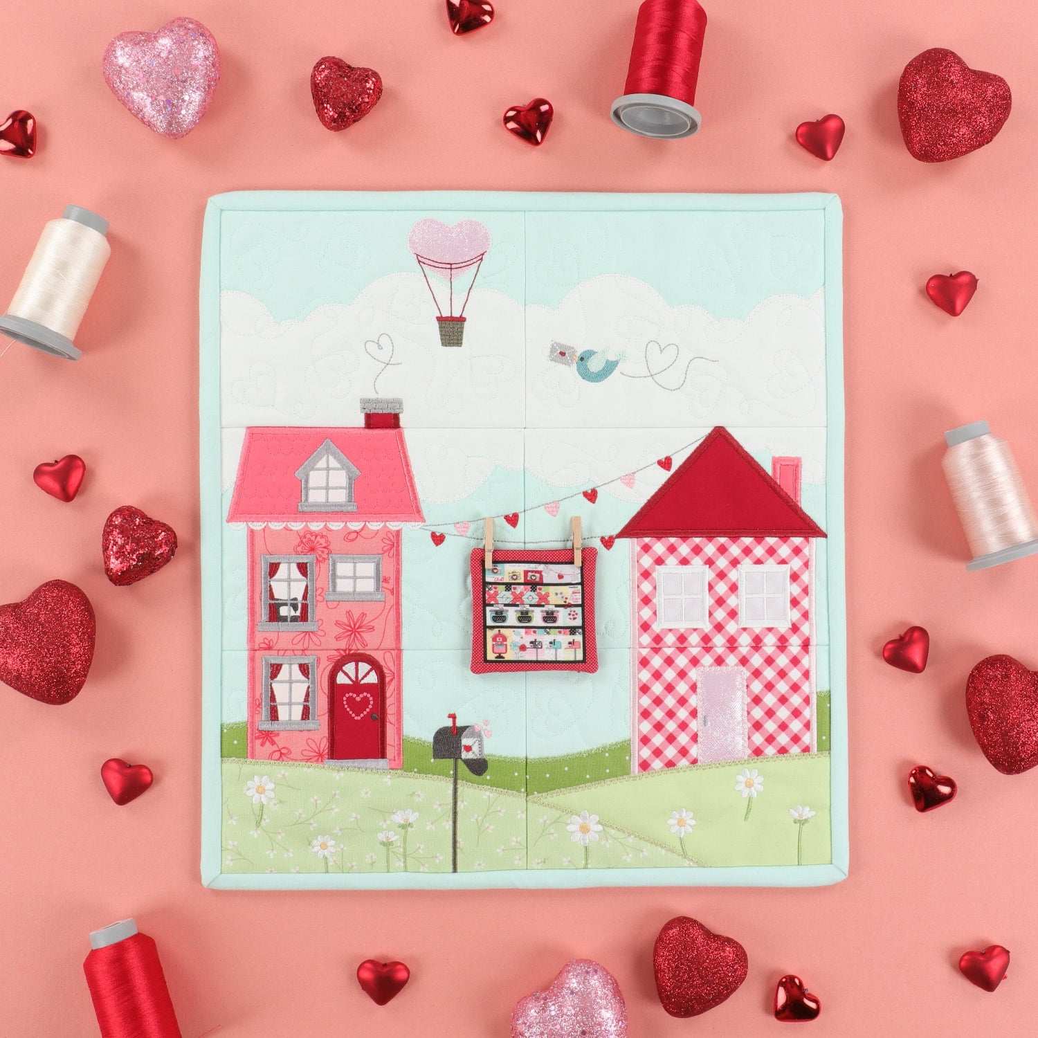 A square quilted fabric panel featuring a whimsical Valentine's Day village scene with two pink houses, a hot air balloon, and a bird, styled on a pink background with red and silver heart decorations and spools of thread.