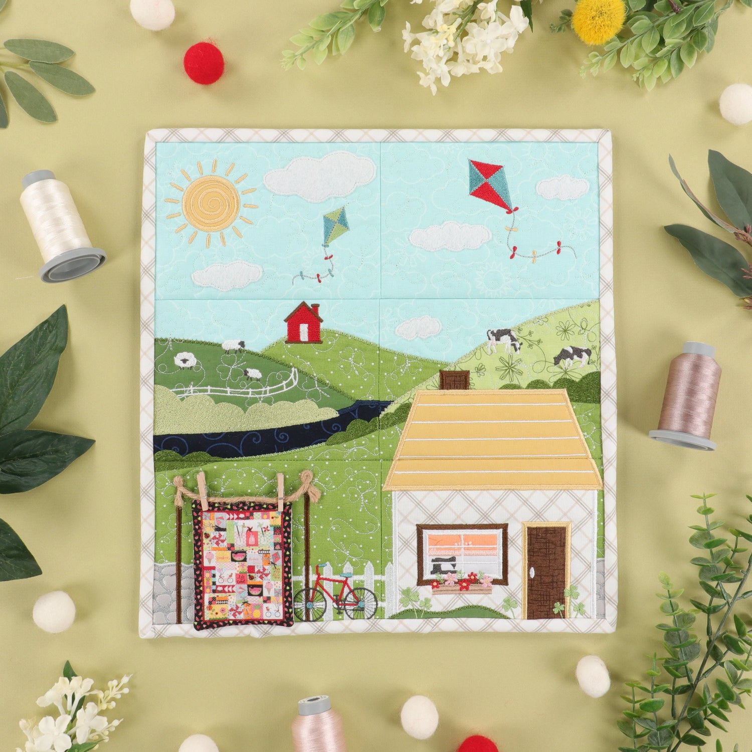 A square quilted fabric panel featuring a whimsical countryside scene with a house, barn, bicycle, flowers, hills, and kites, styled in a flat lay with thread spools, flowers, and leaves on a pale yellow background.