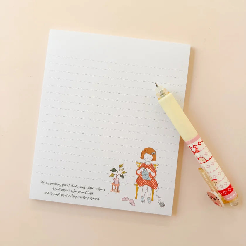 A lined notepad with a cute illustration of a girl sewing and a small poem, styled on a beige background next to a decorative yellow and red patterned pen.