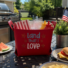 Red cooler with 'Land that I Love' text on a patio table with food and drinks.
