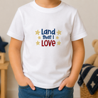 Child wearing a white t-shirt with 'Land that I Love' text and stars, standing in a room with a teddy bear and decorative items.