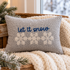 A rectangular gray throw pillow with embroidered snowflakes and the phrase 'let it snow' in blue, styled on a cream knit blanket in a rustic wooden basket with pine branches.