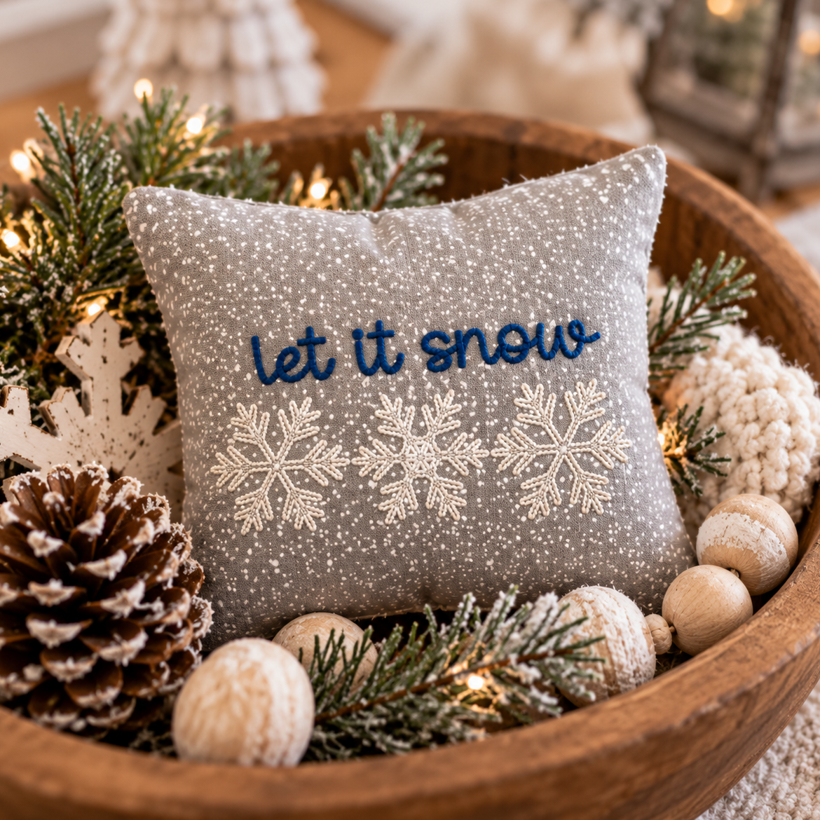 A square gray throw pillow with 'let it snow' embroidered in blue and white snowflakes, nestled in a wooden bowl with pine branches, a pinecone, and holiday decorations.