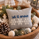 A square gray throw pillow with 'let it snow' embroidered in blue and white snowflakes, nestled in a wooden bowl with pine branches, a pinecone, and holiday decorations.