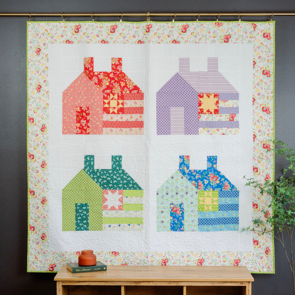 Quilt with colorful house patterns hanging on a wall above a wooden bench.
