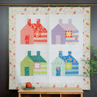 Quilt with colorful house patterns hanging on a wall above a wooden bench.
