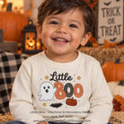 A smiling toddler wearing a cream long-sleeve shirt with a 'Little Boo' Halloween machine embroidery design featuring a ghost, orange and black BOO letters, and stars. The background is a cozy, decorated autumn scene with pumpkins, a lantern, and a 'Trick or Treat' sign.