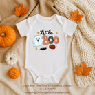 A white short-sleeve baby bodysuit laid flat on a beige fabric background with autumn props, including orange leaves, small pumpkins, and a chunky cream knit blanket. The bodysuit has a machine-embroidered Halloween graphic reading 'Little Boo' with a ghost, stars, and candy.