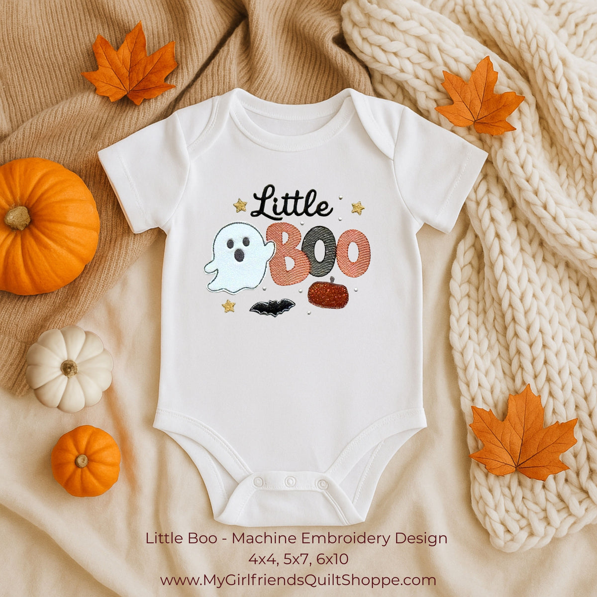 A white short-sleeve baby bodysuit laid flat on a beige fabric background with autumn props, including orange leaves, small pumpkins, and a chunky cream knit blanket. The bodysuit has a machine-embroidered Halloween graphic reading 'Little Boo' with a ghost, stars, and candy.