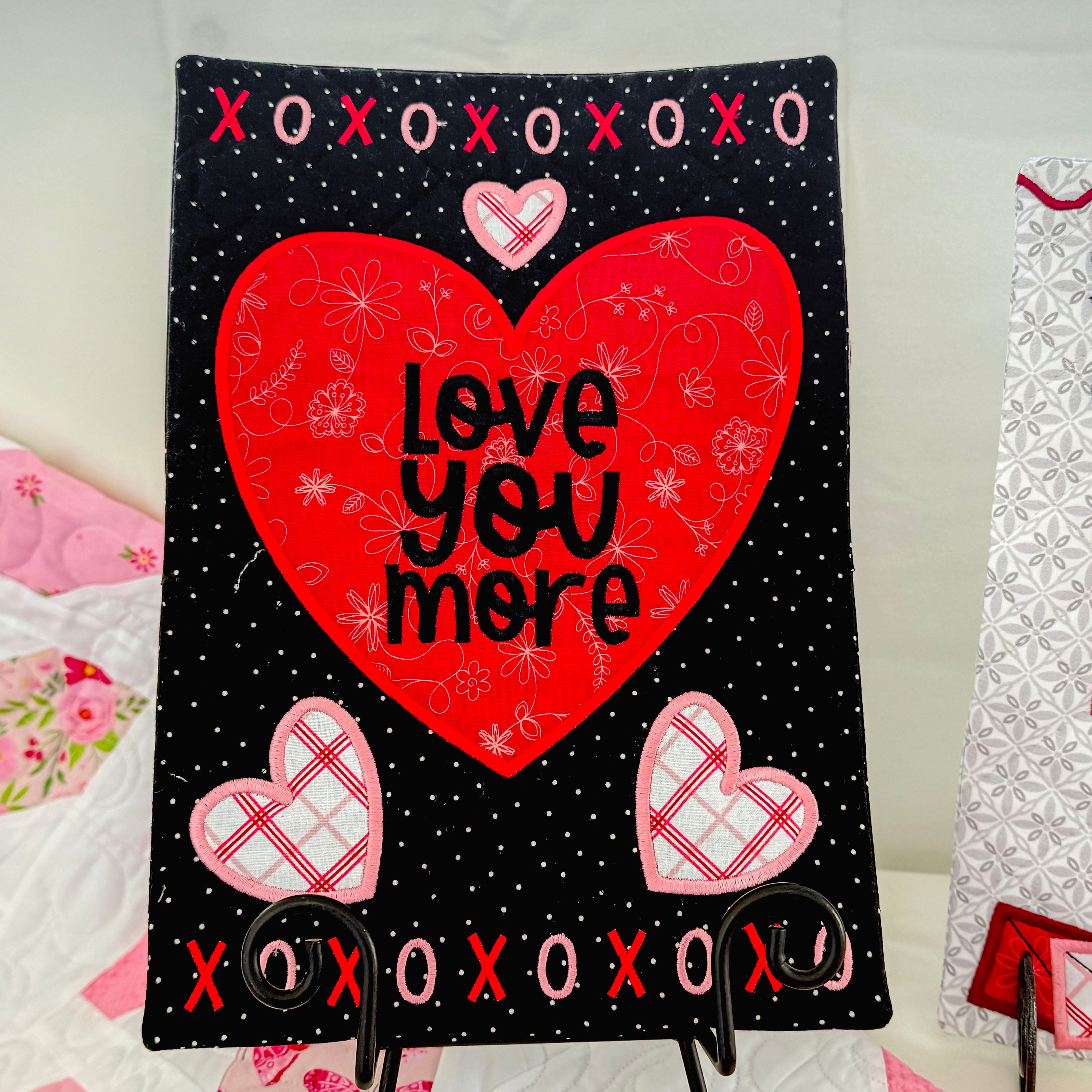 Decorative sign with a red heart and 'Love You More' text on a black background with pink hearts and 'XOXO' text, displayed on an easel.