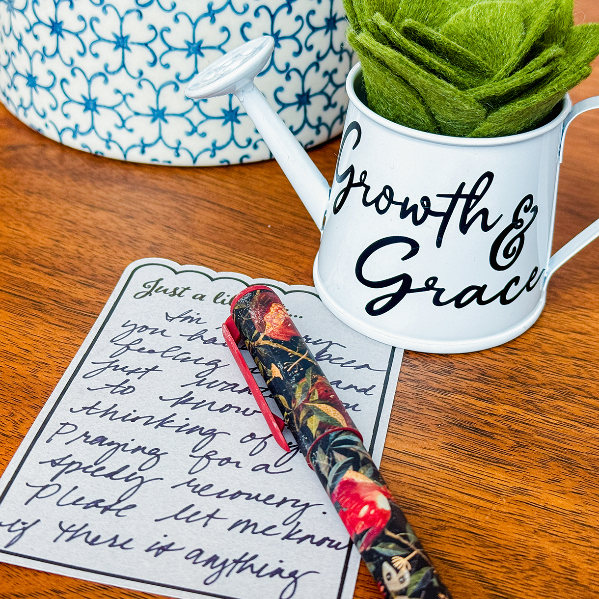 White watering can with 'Growth & Grace' text, floral pen, and handwritten note on a wooden surface.