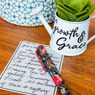 White watering can with 'Growth & Grace' text, floral pen, and handwritten note on a wooden surface.