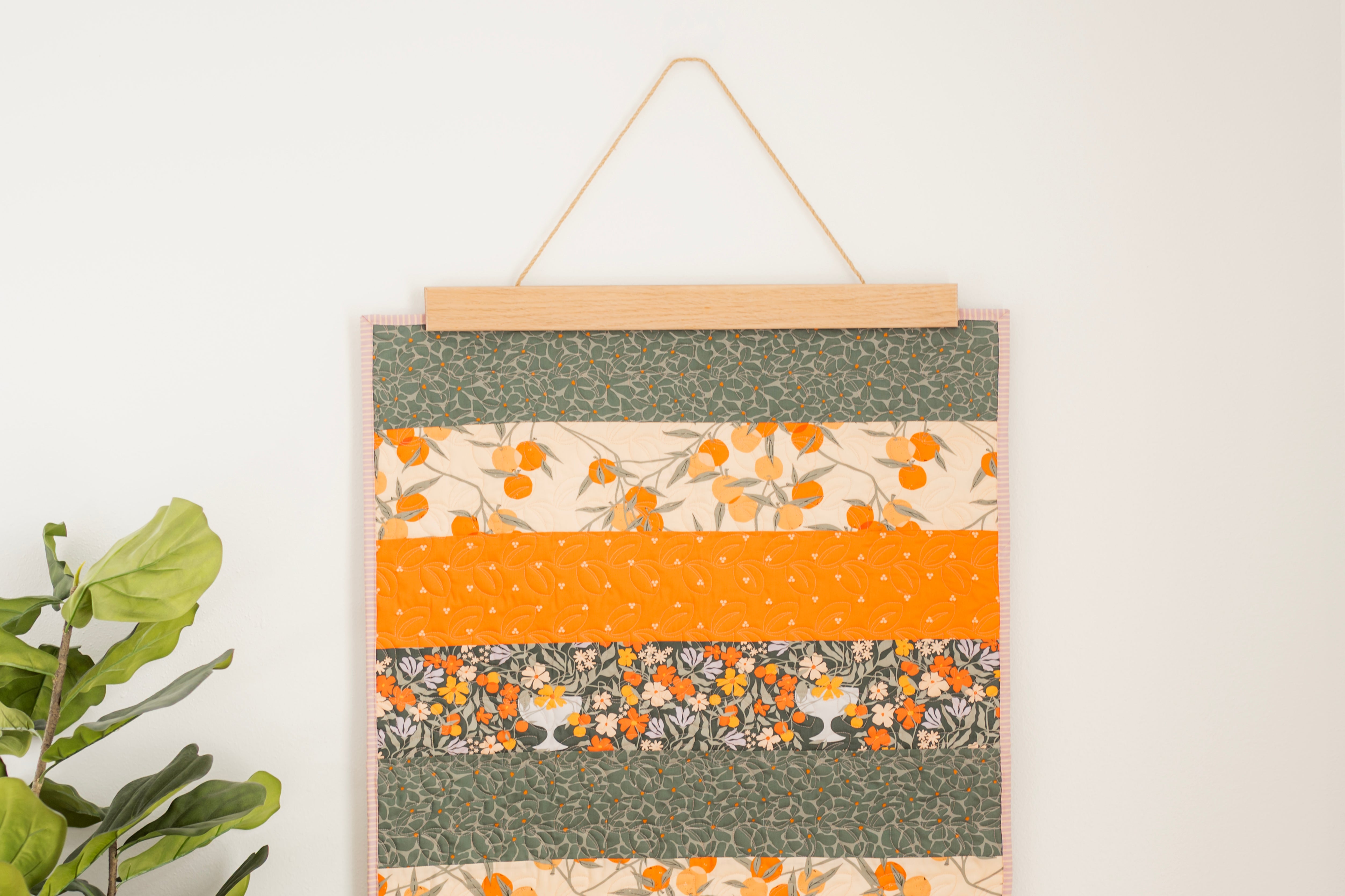 Fabric panel with floral and polka dot patterns in green, orange, and beige hanging on a white wall. 24" wall hanger
