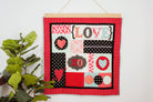 Decorative wall hanging with 'LOVE' and heart designs on a red background, hanging on a white wall with a plant to the left. 36" wall hanger