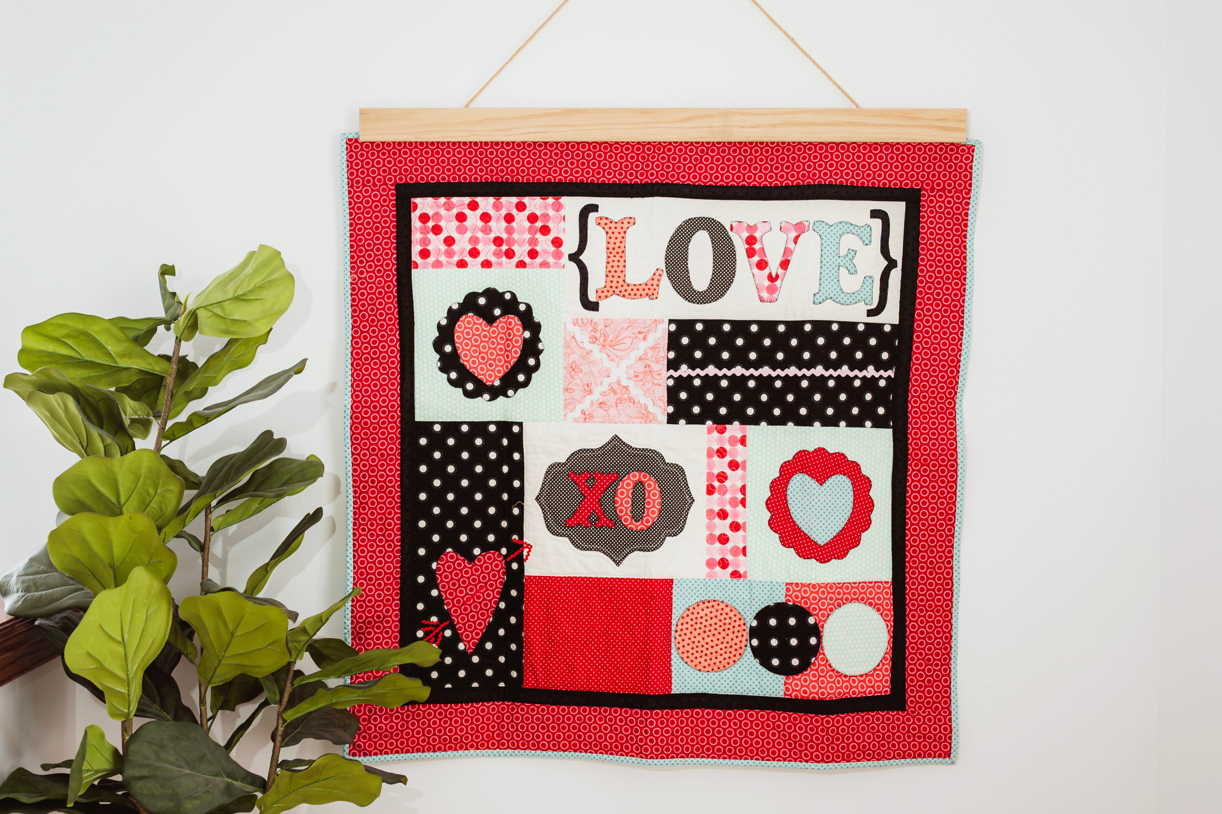 Decorative wall hanging with 'LOVE' and heart designs on a red background, hanging on a white wall with a plant to the left. 36" wall hanger