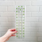 A clear acrylic quilting ruler with green markings and a white brand label, held by a hand against a white painted brick wall.
