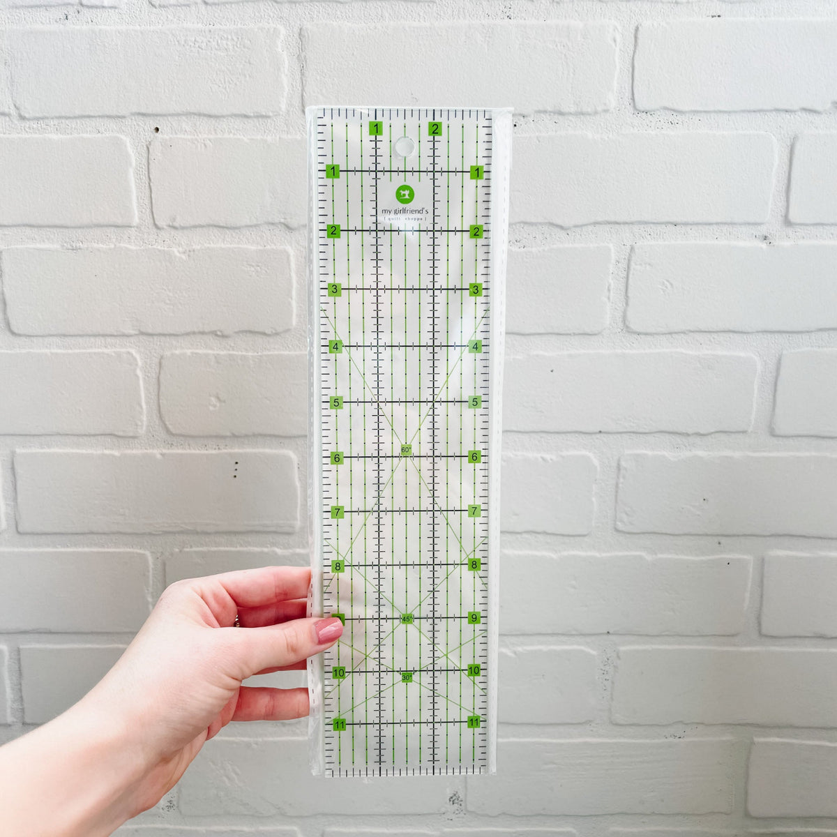 A clear acrylic quilting ruler with green markings and a white brand label, held by a hand against a white painted brick wall.