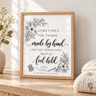 A framed typographic wall art print with black floral illustrations and the phrase “Sometimes the things made by hand are the things that help us feel held,” displayed on a wooden surface beside a vase of dried flowers and folded knit blankets.