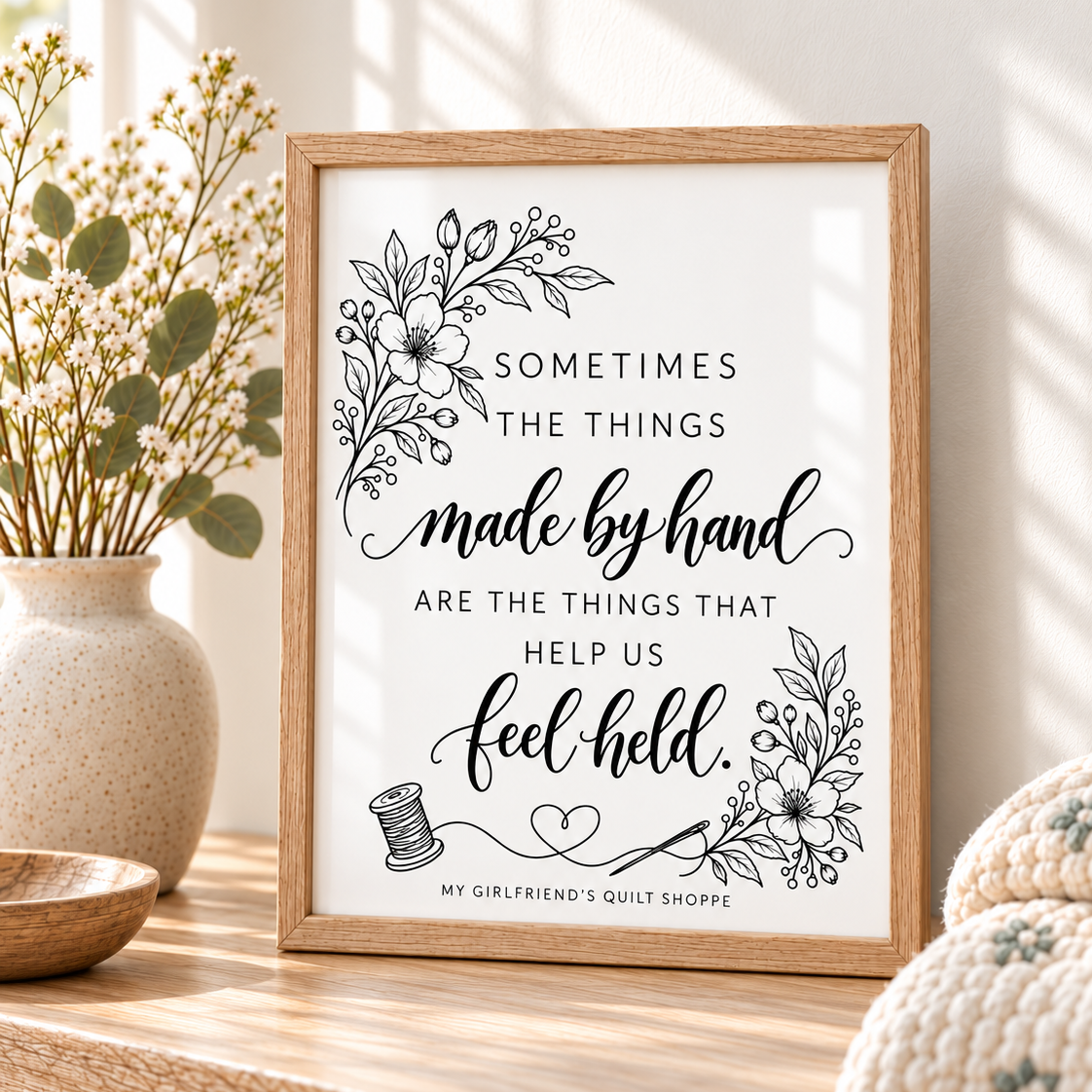 A framed typographic art print with floral line art and the quote “SOMETIMES THE THINGS made by hand ARE THE THINGS THAT HELP US feel held.” The print is displayed on a wooden surface in a warm, styled interior setting with a vase and knit textile nearby.