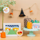A styled tabletop display featuring autumn-themed fabric crafts and decorations, including a small potted plant, a framed quilted mountain landscape, a witch hat hanging on a string, fabric pumpkins, and a decorative sign on a pedestal against a beige wall.