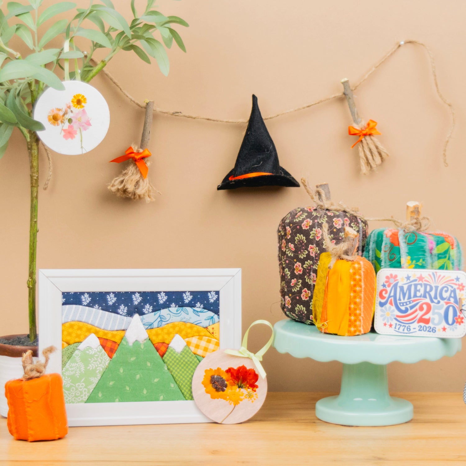 A styled tabletop display featuring autumn-themed fabric crafts and decorations, including a small potted plant, a framed quilted mountain landscape, a witch hat hanging on a string, fabric pumpkins, and a decorative sign on a pedestal against a beige wall.