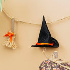 A black felt witch hat with a bright orange band hangs from a jute garland strung across a beige background, with small broom and pumpkin decorations below.