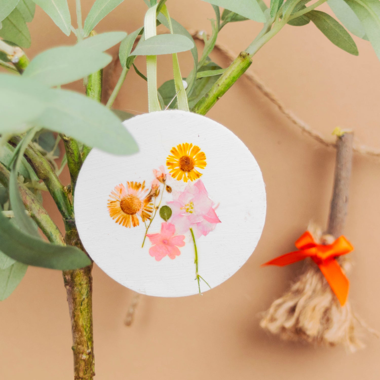 A round white hanging ornament featuring pressed flowers, displayed on a green plant against a warm beige background with a small decorative broom hanging nearby.
