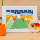 A framed handmade quilted mountain landscape art piece with colorful fabric patterns, displayed on a wooden surface next to a small orange fabric pumpkin and a round floral ornament.
