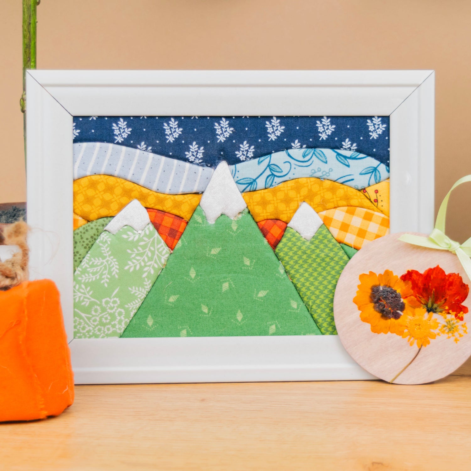 A framed handmade quilted mountain landscape art piece with colorful fabric patterns, displayed on a wooden surface next to a small orange fabric pumpkin and a round floral ornament.