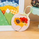 A round wooden hanging ornament with pressed yellow and red flowers, tied with a pale green ribbon, leaning on a wooden surface next to a quilted frame and a mint cake stand.