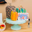 A styled tabletop display featuring several fabric-covered decorative pumpkins and a small round fabric ornament, arranged on a pale blue pedestal against a beige wall. A framed quilt and a small black hat hang in the background, and a decorative tag reads "America 2026.