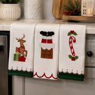 Three white kitchen towels with Christmas-themed embroidery hanging on a countertop. The left towel shows a reindeer with green and red gift boxes, the middle towel shows Santa's boots and chimney, and the right towel shows a candy cane with holly.